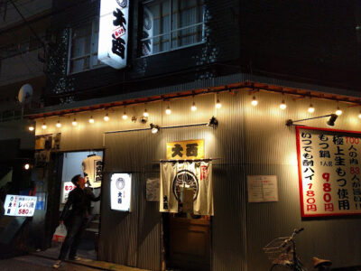 夜の居酒屋。暖色系の提灯と電球が灯り、賑やかな雰囲気。看板には「大吉」と書かれている。