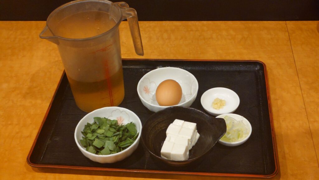 料理の材料がトレイに並べられている。計量カップ、豆腐、卵、刻み野菜、薬味などが見える。